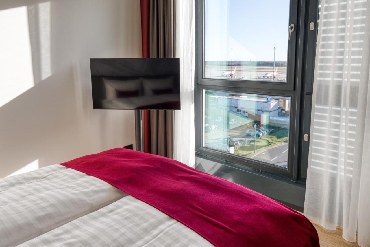 Intercityhotel Berlin Airport Ber Terminal 1 & 2 Schönefeld Extérieur photo The photo shows a modern hotel room with a large bed covered in white linens and a burgundy throw blanket. In the background, there is a large window that lets in natural light, offering a view of the outside. A flat-screen TV is mounted on the wall 