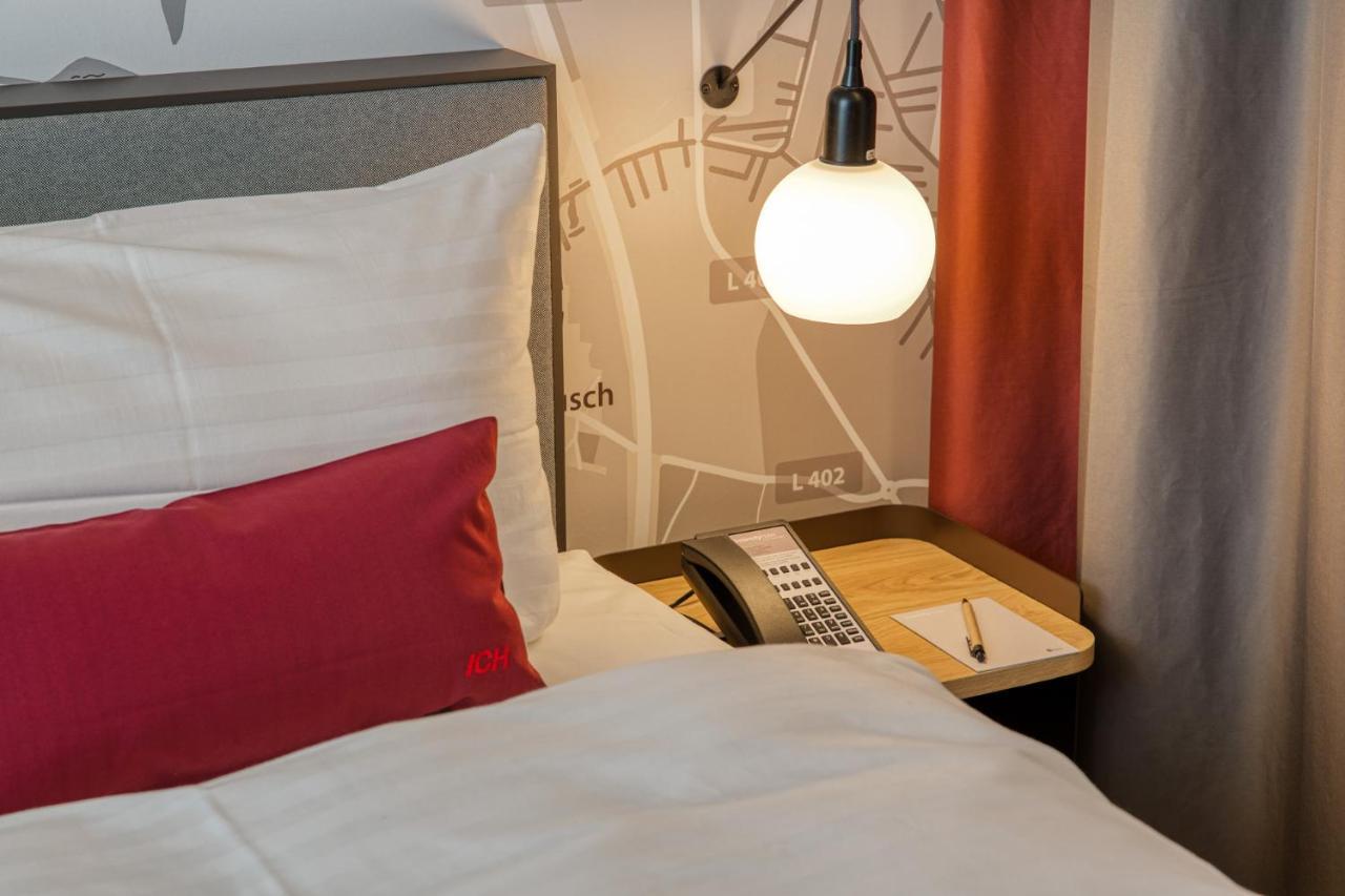 Intercityhotel Berlin Airport Ber Terminal 1 & 2 Schönefeld Extérieur photo The photo shows a cozy bedroom setup. There's a neatly made bed with white bedding and a red decorative pillow. Beside the bed, a small bedside table holds a telephone and a notebook or notepad, along with a pen. A round pendant light hangs above the
