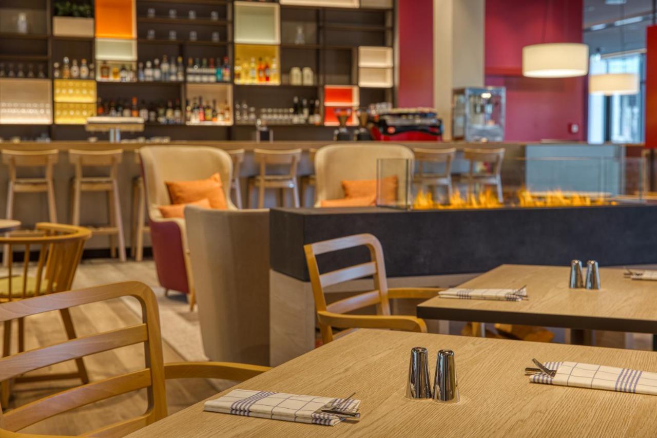 Intercityhotel Berlin Airport Ber Terminal 1 & 2 Schönefeld Extérieur photo The photo shows a modern restaurant or bar interior. In the foreground, there are wooden tables set with napkins and condiments. The seating includes wooden chairs and plush seating in various colors in the background. A stylish bar area is visible w